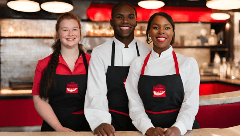 Three Game Day Grill employees are pictured. They are smiling and are wearing black aprons, on top of white or red shirts.