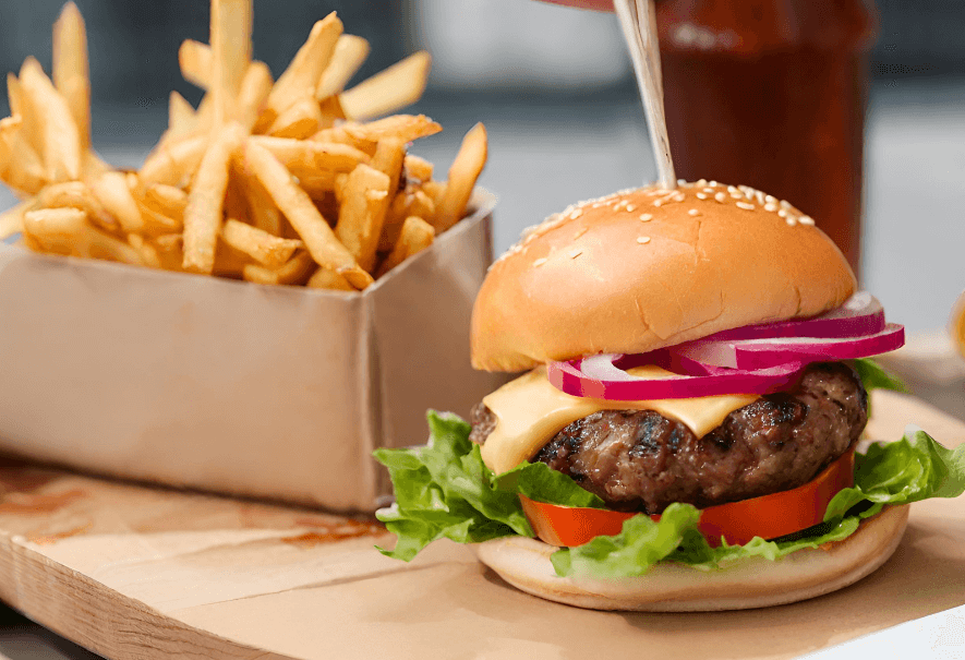 A burger with a sesame seed bun, onions, cheese, tomato, and lettuce is pictured. The burger has a stick in the middle of it. To the right of the burger there is a box of fries.